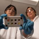 Two researchers stand in a lab looking at a photocell, wearing PPE